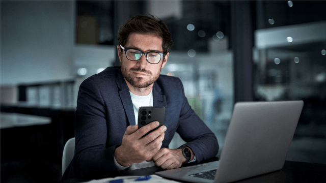Mann in Anzug mit Brille, der auf sein Smartphone schaut, während er an einem Schreibtisch mit Laptop arbeitet.