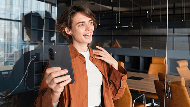 Junge Frau in modernem Büro mit Smartphone, lächelnd und gestikulierend, symbolisiert innovative Kommunikation der Brodos AG.