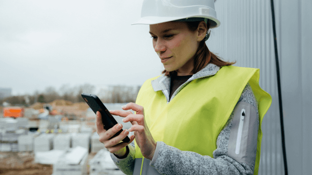 Frau in Sicherheitskleidung und Helm nutzt ein Smartphone auf einer Baustelle, um Informationen zum Samsung Galaxy Tab S10 Lite EE abzurufen.